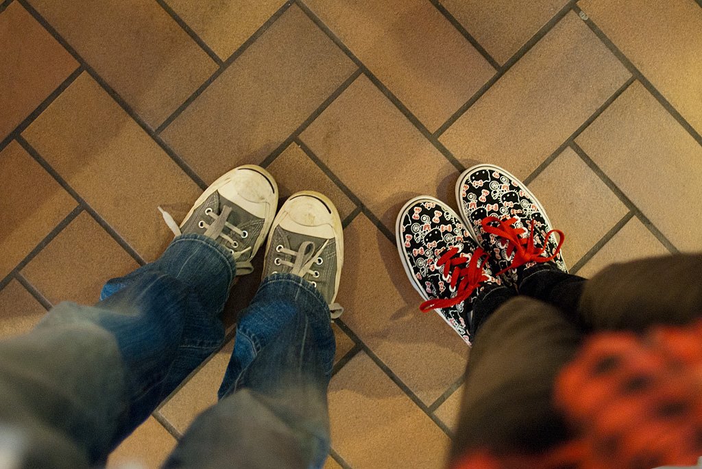 Andy and Michelle's shoes from a top-down view, in 2011. Andy is wearing slate blue Converses, while Michelle is wearing Hello Kitty Vans, on a brown tiled floor.