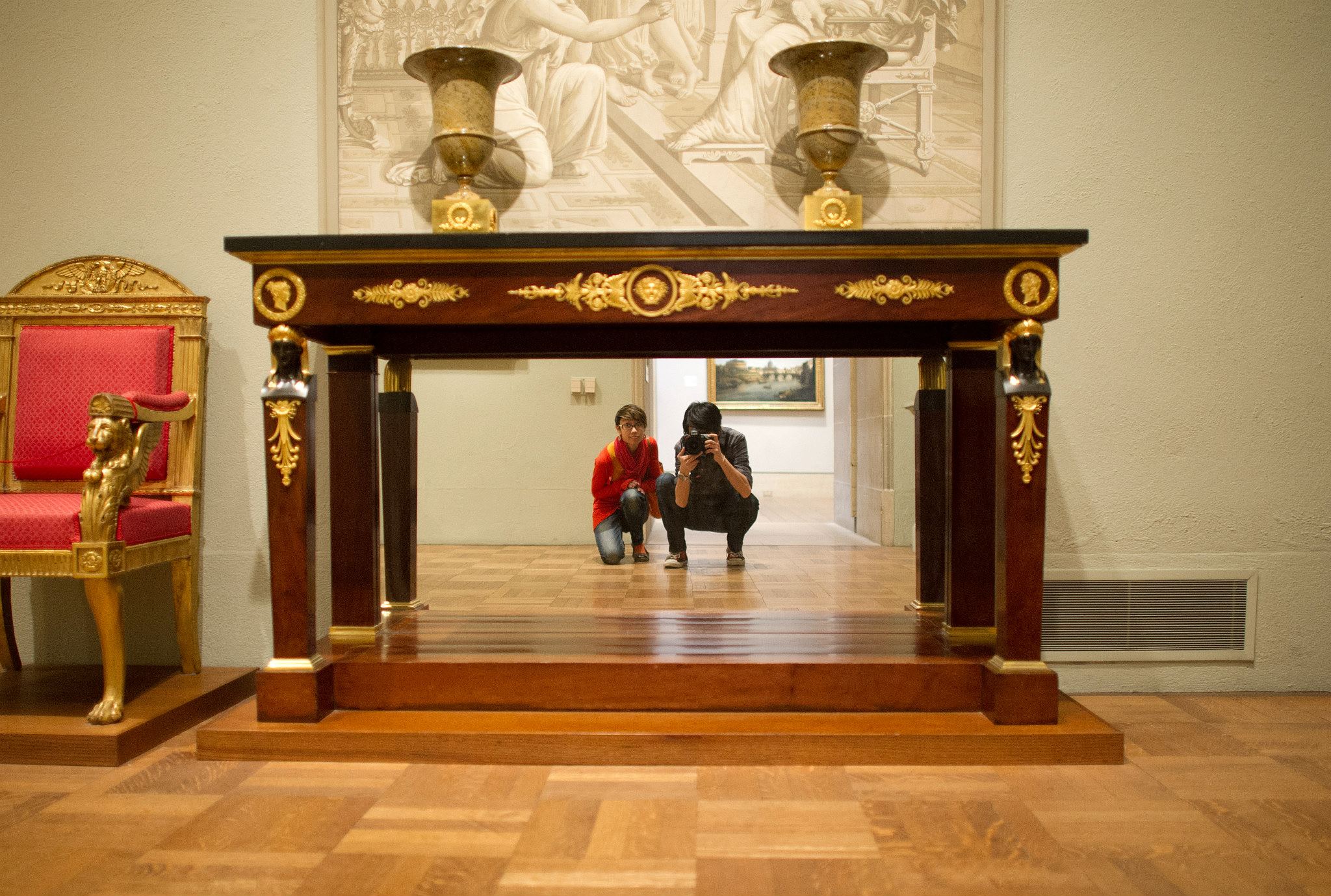 Andy and Michelle are in a museum. They are kneeling, looking under a wooden mantel decorated with gold flourishes, where there is a mirror. Michelle has a pixie cut and red sweater, while Andy is wearing a black and gray checkered button-down shirt, taking a photo of both of them in the mirror. He is holding a Nikon DSLR.