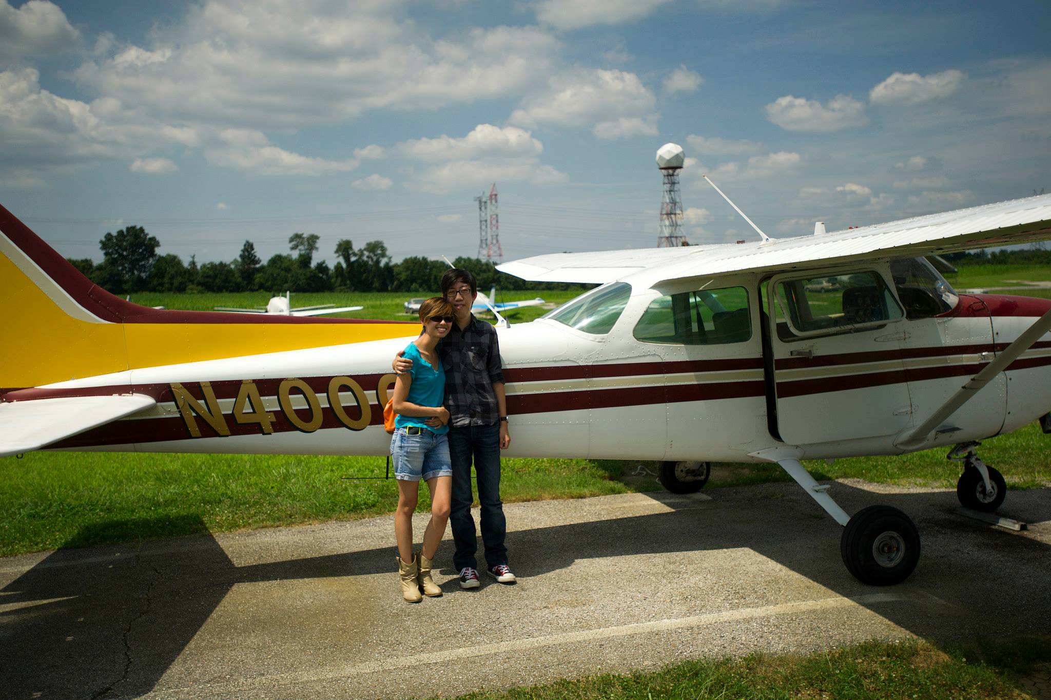 Andy and Michelle are standing in front of a Cessna plane that Andy got trained on. Behind it is a field of green grass and blue skies. Michelle wears a blue v-neck shirt, jean shorts, and brown cowboy boots. Andy wears a black and gray checkered button-up shirt, over blue jeans, and red Converses.