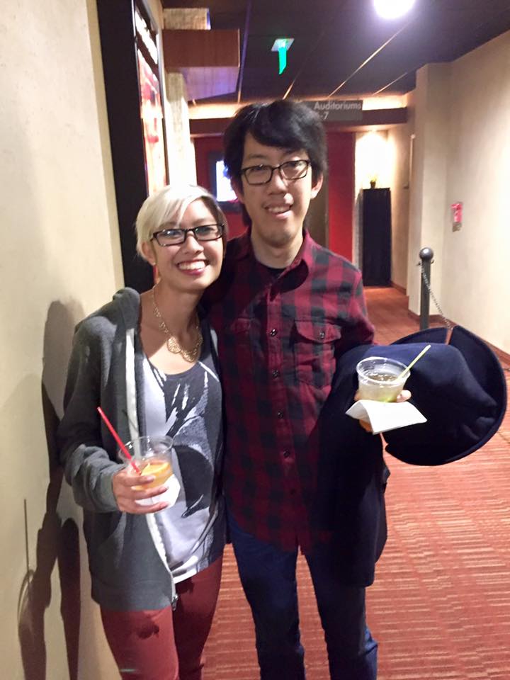 Full body image of Michelle and Andy waiting in a movie theater. They are smiling and hugging each other, holding alcoholic drinks. Michelle has short blonde pixie cut hair, wearing a gray hoodie over a Darth Vader tank top, over red jeans. Andy is wearing a red and black checkered button down shirt with his sleeves rolled up, over some blue jeans.