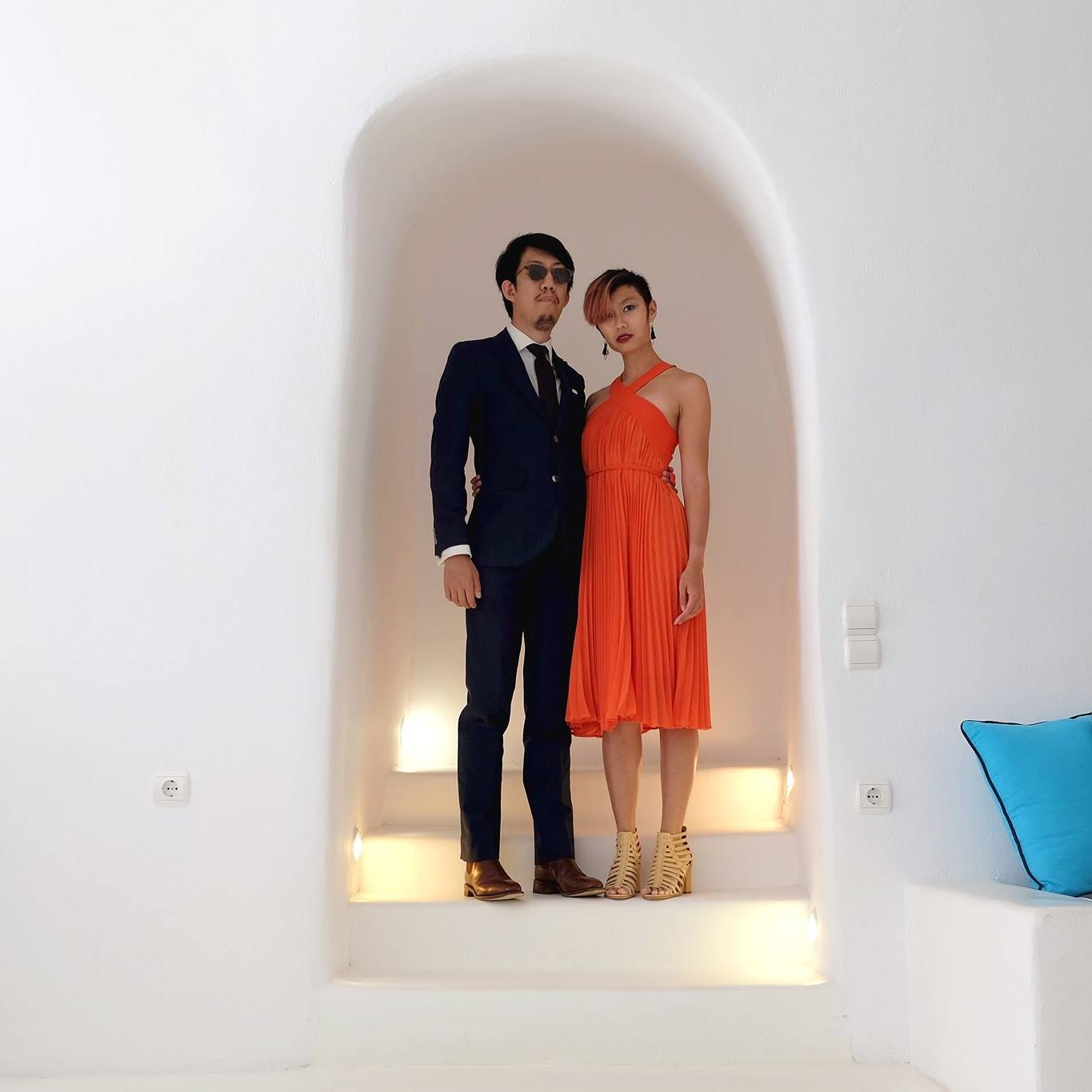 Andy and Michelle stand in a white alcove in Santorini, Greece. Andy is wearing a navy blue suit, and sunglasses, while Michelle is wearing a pleated orange halter dress, both looking off into the distance.