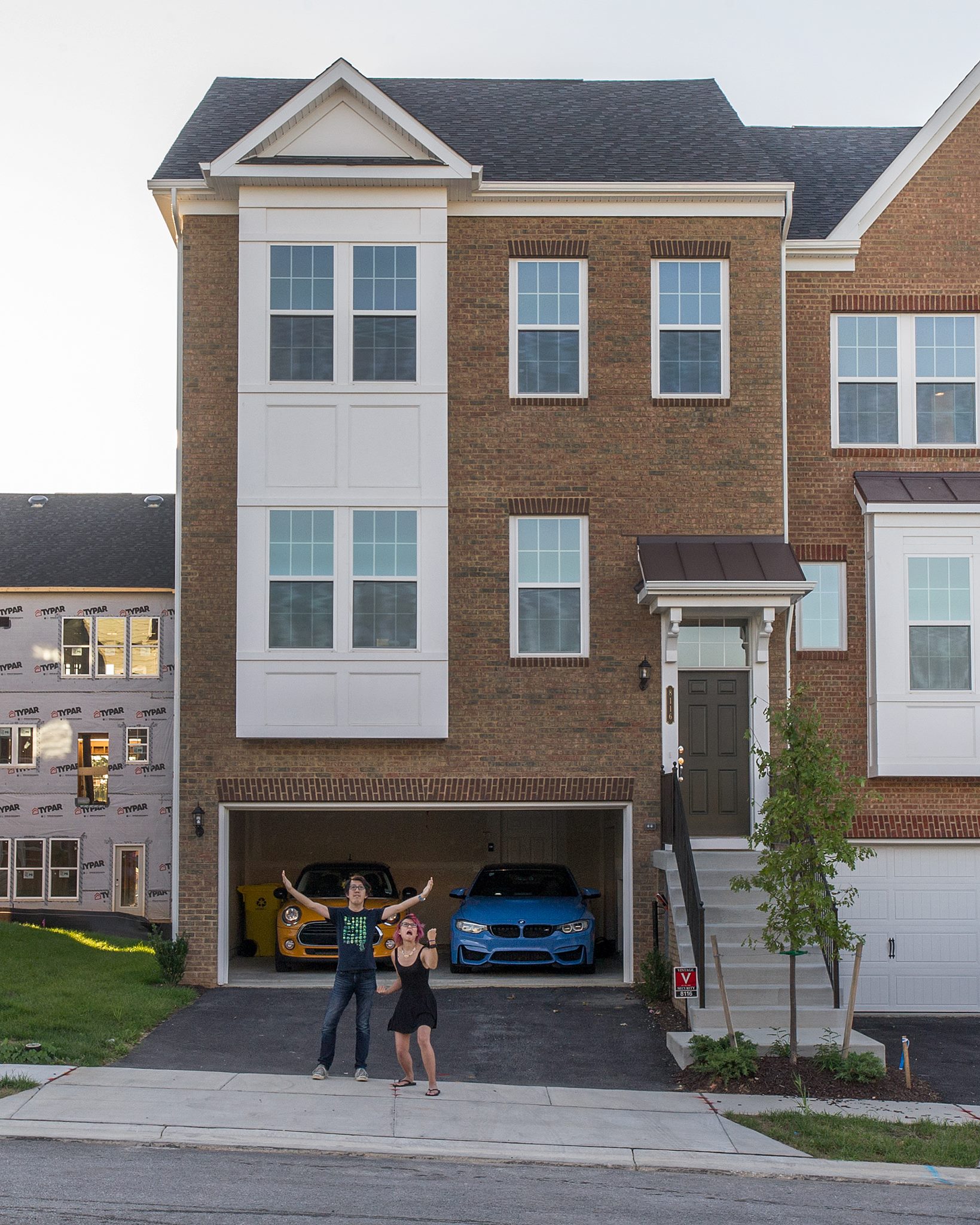 Front view of Michelle and Andy's townhome in Maryland, taken shortly after they moved in. Andy has his arms wide open above his head, and Michelle is pumping her fist and mid-scream. The facade is a brown and red brick, with white bay windows on two floors. The garage door is open, showing Michelle's previous Mini Cooper in orange, and Andy's current BMW M4 in a bright blue.