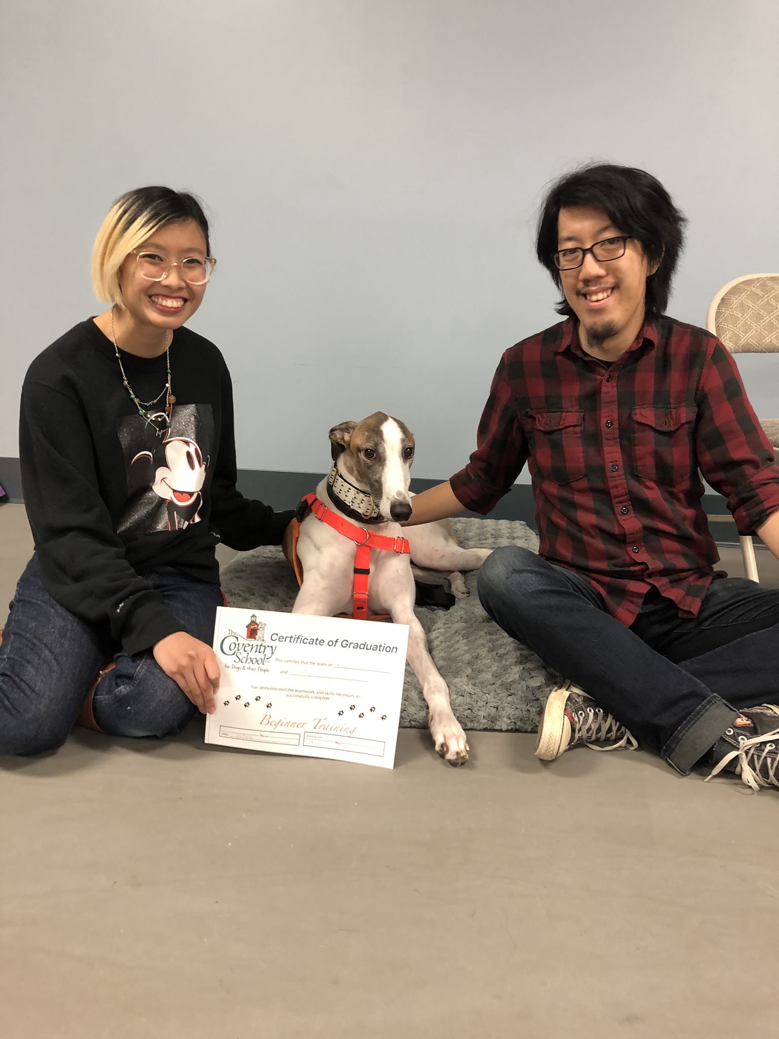 Michelle and Andy sit on the floor with their dog, Fig, laying between them. Fig is a greyhound with brown markings over his eyes, with a mostly white body, with occasional brown markings. In front of Fig, Michelle holds a certificate, saying 'Certificate of Graduation', signifying that Fig has graduated his dog training class.