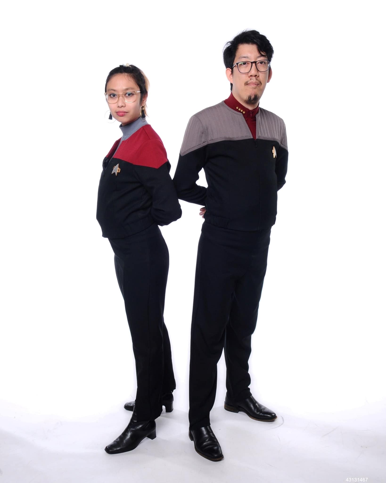 Photo of Michelle and Andy in their Star Trek cosplay. Michelle is wearing a red and black Voyager-era captain's uniform over a slate blue turtleneck, and with black heeled boots. Andy is wearing a Deep Space 9 era captains uniform, gray and black over a red shirt, and with black boots. They stand with their hands behind their backs in military fashion.